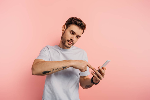 αυτοπεποίθηση νεαρός άνδρας δείχνοντας με το δάχτυλο στο smartphone κατά τη διάρκεια της κλήσης βίντεο σε ροζ φόντο - Φωτογραφία, εικόνα