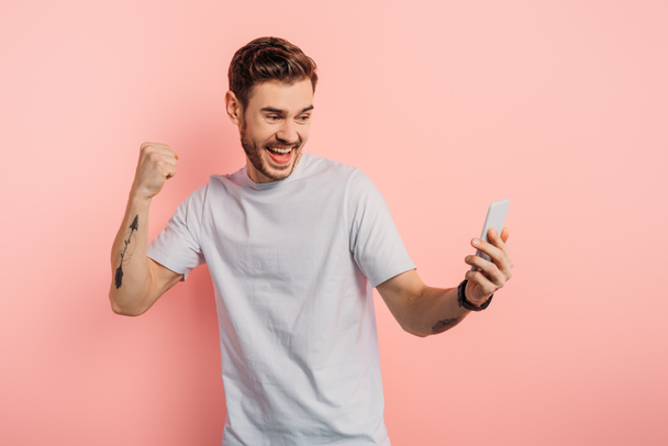 vzrušený mladý muž ukazuje vítězný gesto během videohovoru na smartphone na růžovém pozadí - Fotografie, Obrázek