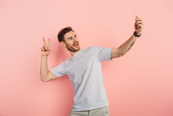 excited young man showing victory gesture while taking selfie on smartphone on pink background - Fotografie, Obrázek