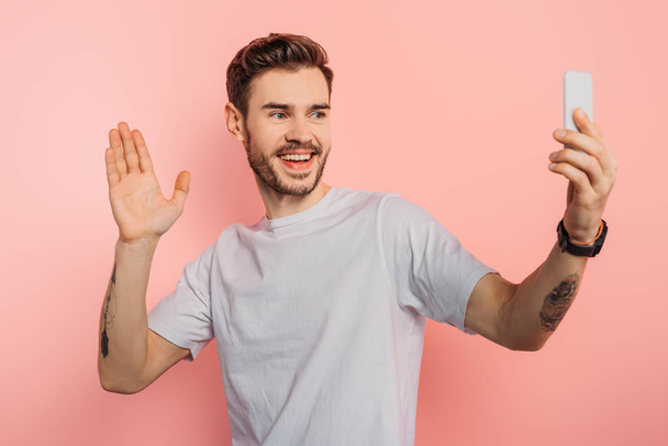 excited young man waving hand while having video chat on smartphone on pink background - Фото, зображення