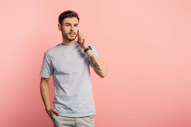 serious handsome man talking on smartphone while holding hand in pocket on pink background - Zdjęcie, obraz