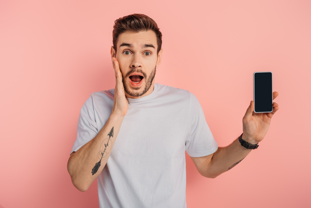 shocked man touching face while showing smartphone with blank screen on pink background - Foto, Imagen