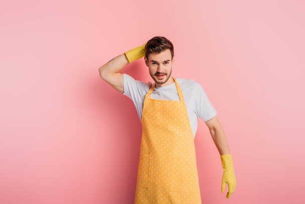 smiling young man in apron and rubber gloves touching head while looking at camera on pink background - Photo, Image