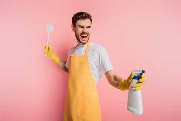 angry young man in apron and rubber gloves holding spray bottle and plunger on pink background - Photo, Image