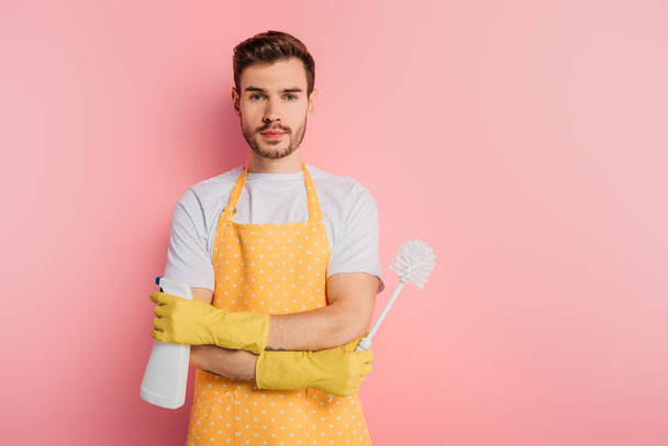serious young man in apron and rubber gloves holding spray bottle and toilet brush on pink background - Foto, imagen