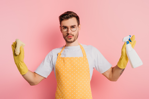 serious young man in apron and rubber gloves holding spray bottle and rag on pink background - Foto, afbeelding