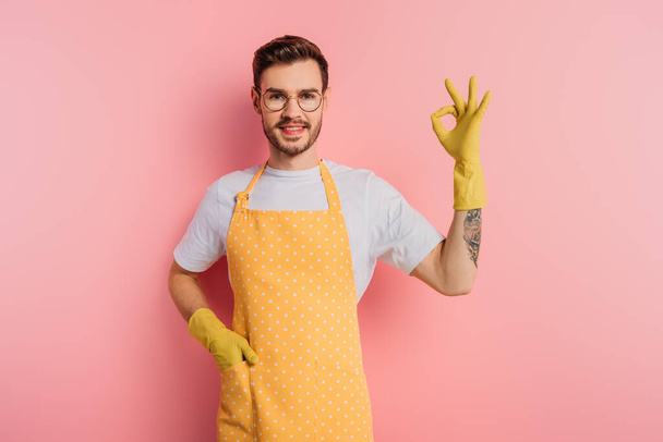happy young man in apron and rubber gloves showing okay gesture on pink background - Fotografie, Obrázek