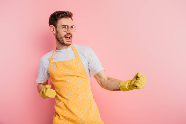 excited young man in apron and rubber gloves showing winner gesture on pink background - Valokuva, kuva