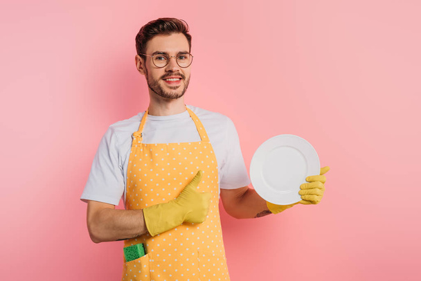 χαρούμενος νεαρός άνδρας με ποδιά και λαστιχένια γάντια κρατώντας καθαρό πιάτο και δείχνοντας τον αντίχειρα επάνω σε ροζ φόντο - Φωτογραφία, εικόνα