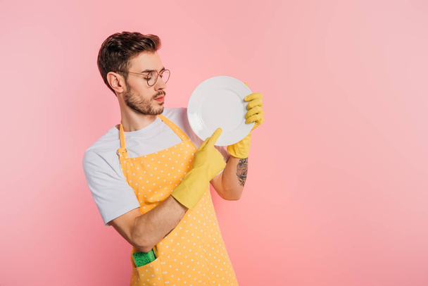 serious young man in apron and rubber gloves touching clean plate on pink background - Foto, Imagem