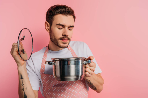 pleased young man in apron enjoying flavor with closed eyes while opening saucepan on pink background  - Foto, afbeelding