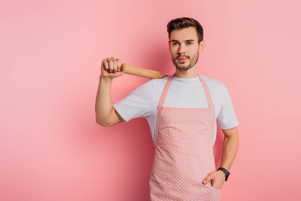 cheerful young man in apron holding rolling pin while standing with hand in pocket on pink background - Фото, изображение