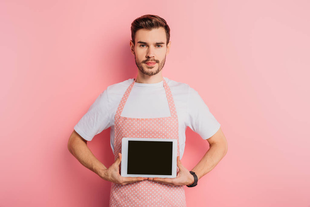 serious young man in apron showing digital tablet with blank screen on pink background - Fotó, kép