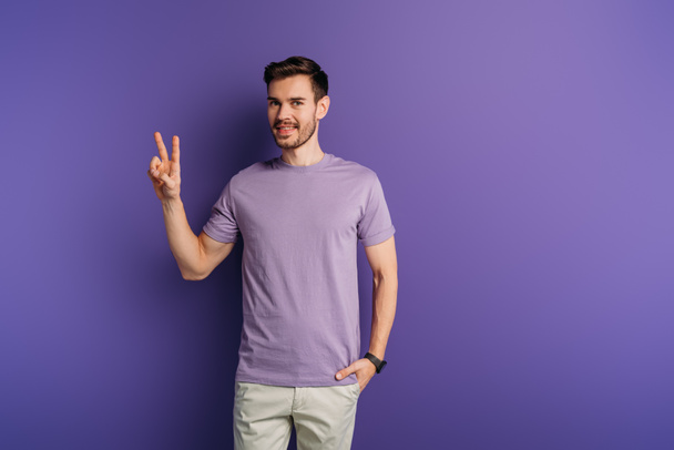 smiling young man showing victory gesture while holding hand in pocket on purple background - Photo, Image