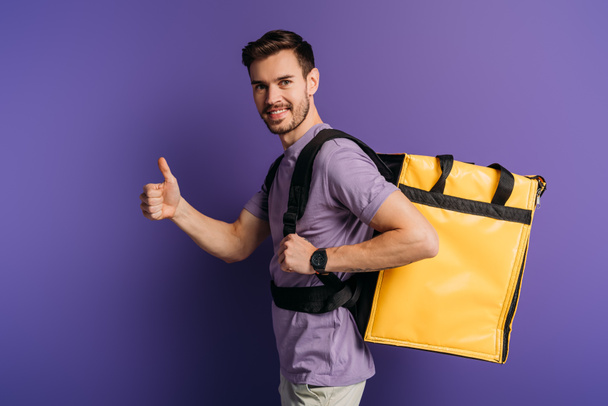 happy delivery man showing thumb up while looking at camera on purple background - Foto, Imagen