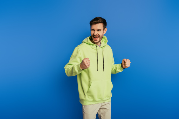 excited young man showing winner gesture while looking away on blue background - Фото, изображение