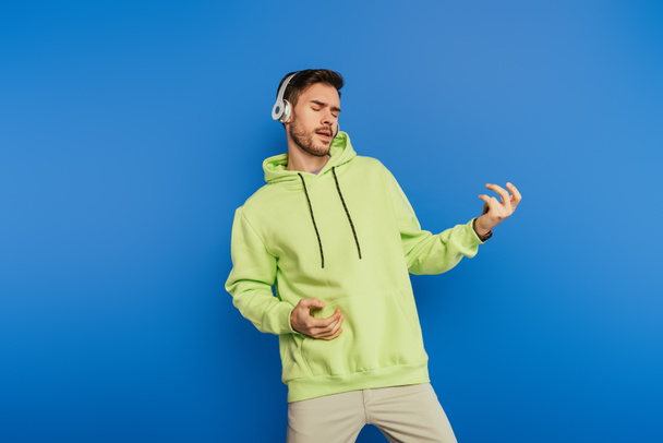 inspired young man in wireless headphones imitating playing guitar isolated on blue - Foto, imagen