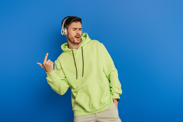 excited young man in wireless headphones singing with closed eyes on blue background - Foto, Imagen