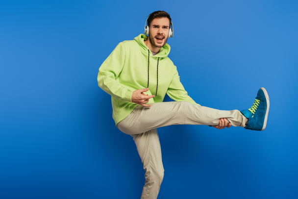 vrolijke jongeman in draadloze hoofdtelefoon dansen en zingen op blauwe achtergrond - Foto, afbeelding