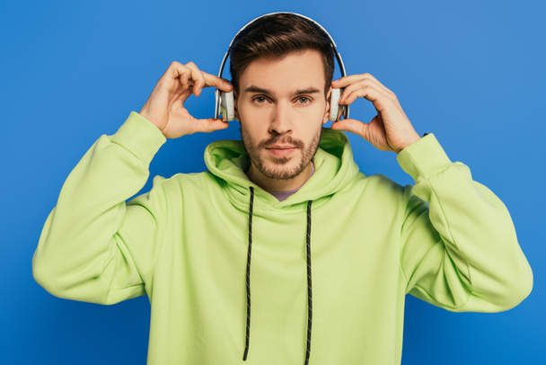 serious young man touching wireless headphones while looking at camera isolated on blue - Photo, Image