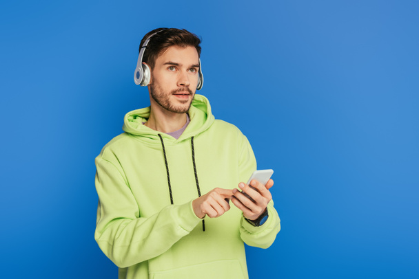 στοχαστικός νεαρός άνδρας με ασύρματα ακουστικά χρησιμοποιώντας smartphone και κοιτάζοντας μακριά απομονώνονται σε μπλε - Φωτογραφία, εικόνα