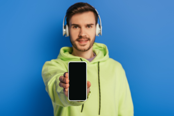 selective focus of cheerful young man in wireless headphones showing smartphone with blank screen on blue background - Photo, Image