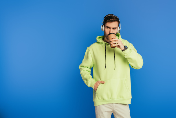 marzycielski młodzieniec w bezprzewodowych słuchawkach pijący kawę i odwracający wzrok odizolowany na niebiesko - Zdjęcie, obraz