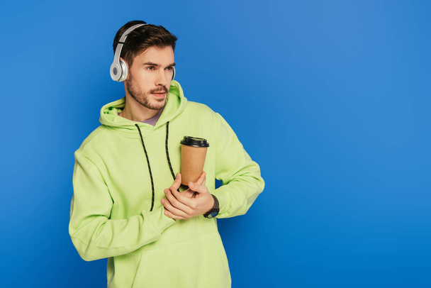 verträumter junger Mann mit drahtlosen Kopfhörern, Kaffee to go in der Hand und isoliert auf blauem Grund schauend - Foto, Bild