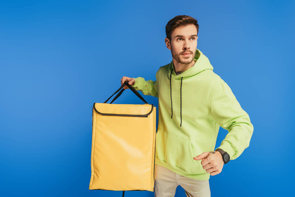 serious delivery man looking away while holding thermo backpack isolated on blue - Foto, Imagem