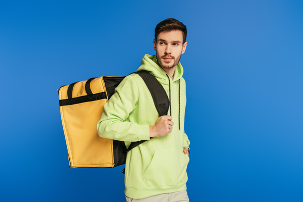 handsome delivery man carrying thermo backpack while looking away isolated on blue - Фото, изображение