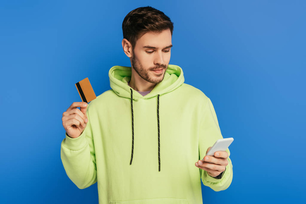 attentive young man using smartphone while holding credit card isolated on blue - Photo, image