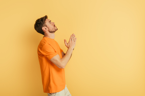 side view of serious young man looking up while praying on yellow background - Photo, image