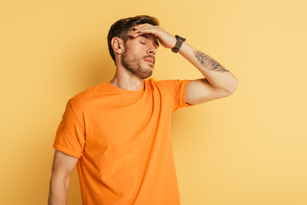 exhausted young man touching forehead while standing with closed eyes on yellow background - Fotografie, Obrázek