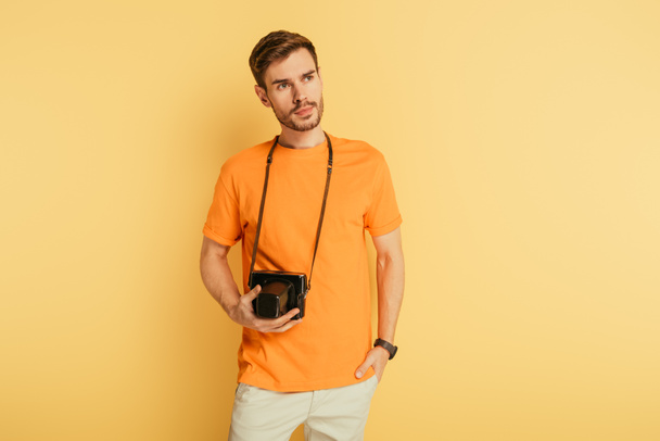 handsome thoughtful photographer with digital camera holding hand in pocket and looking away on yellow background - Фото, изображение