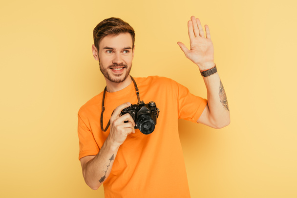 handsome, cheerful photographer with digital camera waving hand on yellow background - Photo, Image