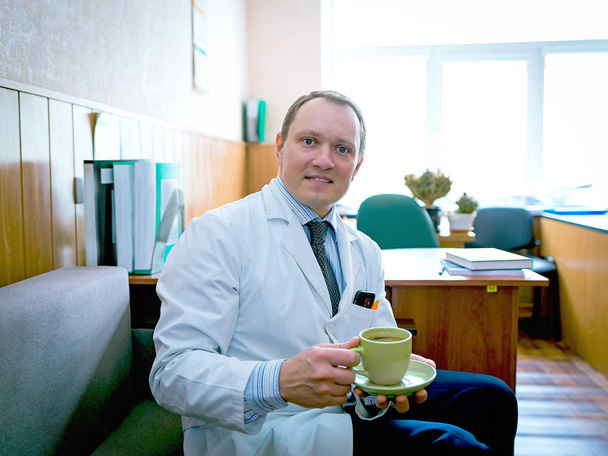 młody atrakcyjny lekarz w białym płaszczu trzyma filiżankę kawy  - Zdjęcie, obraz