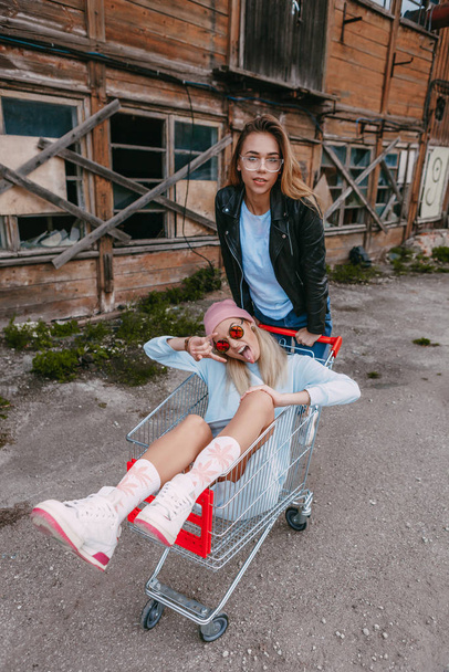 A long haired blonde woman wearing a powder blue dress, pink woollen hat and dark sunglasses showing reflections of the sun, is having great fun pushing her female friend, who is wearing a black leath - Fotografie, Obrázek