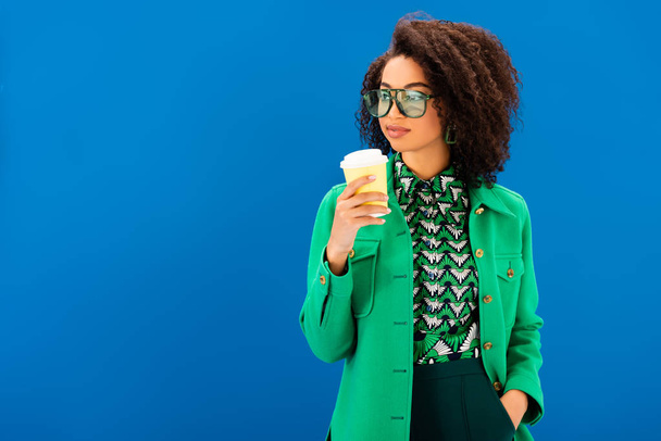 stylish african american woman in jacket holding paper cup isolated on blue - Фото, зображення