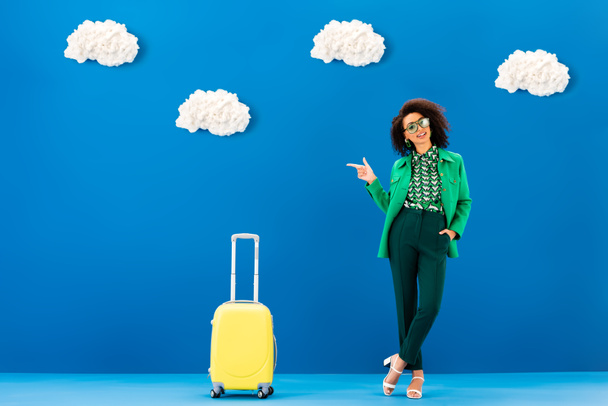 uśmiechnięta Afroamerykanka wskazująca palcem i stojąca w pobliżu torby na niebieskim tle z chmurami  - Zdjęcie, obraz