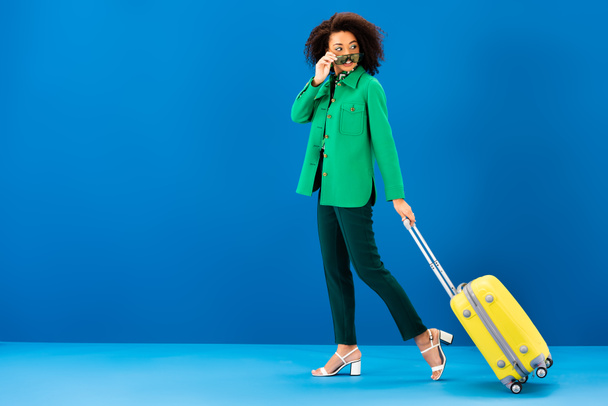 smiling african american woman holding sunglasses and holding travel bag on blue background  - 写真・画像