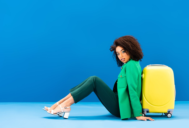 stílusos afro-amerikai nő ül közelében utazási táska kék háttér  - Fotó, kép
