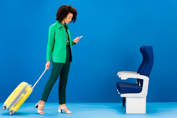 donna afro-americana sorridente che tiene la borsa da viaggio e utilizza lo smartphone su sfondo blu
  - Foto, immagini