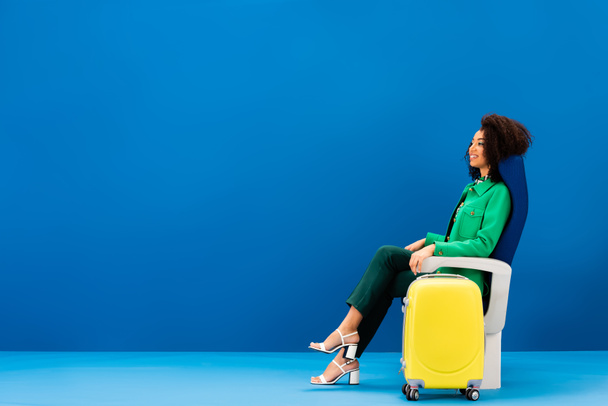 sonriente mujer afroamericana sentada en el asiento cerca de la bolsa de viaje sobre fondo azul
  - Foto, Imagen
