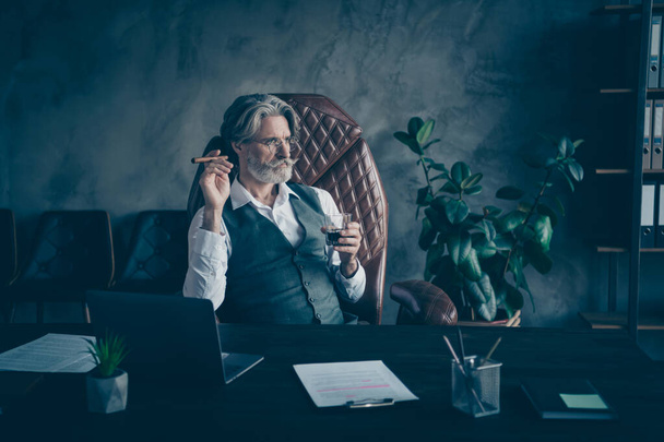 Portrait of his he nice attractive chic classy serious focused gray-haired man drinking liquor smoking cigar inspiration at modern loft industrial style interior work place station indoors - Foto, Imagen