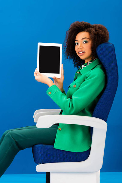smiling african american sitting on seat and holding digital tablet on blue background  - Photo, Image