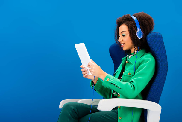 zijaanzicht van glimlachend Afrikaans Amerikaans met behulp van digitale tablet en het luisteren naar muziek geïsoleerd op blauw  - Foto, afbeelding