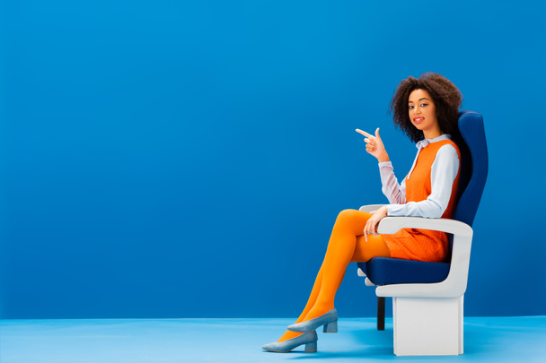 sonriente afroamericano en vestido retro sentado en el asiento y señalando con el dedo sobre fondo azul
  - Foto, Imagen