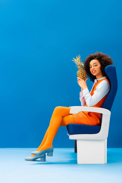 smiling african american in retro dress sitting on seat and holding pineapple on blue background  - Photo, image
