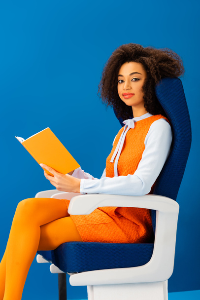 усміхнений афроамериканський американець у ретро-сукні тримає книгу і сидить на сидінні ізольовано на синьому
  - Фото, зображення