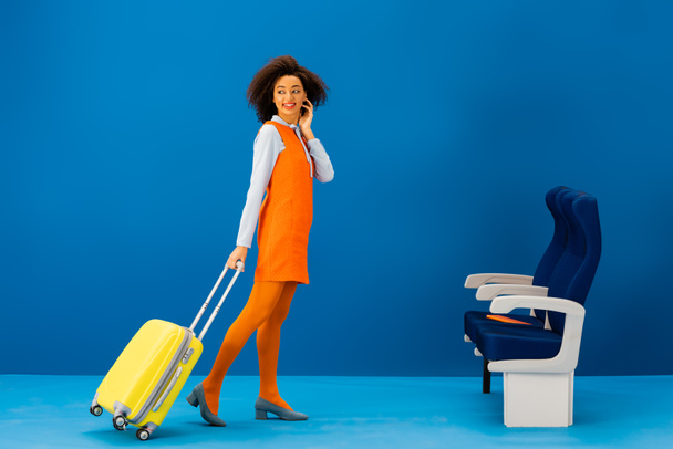 sonriente afroamericano en vestido retro caminando con bolsa de viaje sobre fondo azul
  - Foto, Imagen